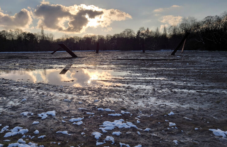 View of a ploughed field with an artwork in the middle, consisting of 7 giant steel slabs positioned in various angles. The sun behind a cloud if reflected in the pool of water in front of the artwork.